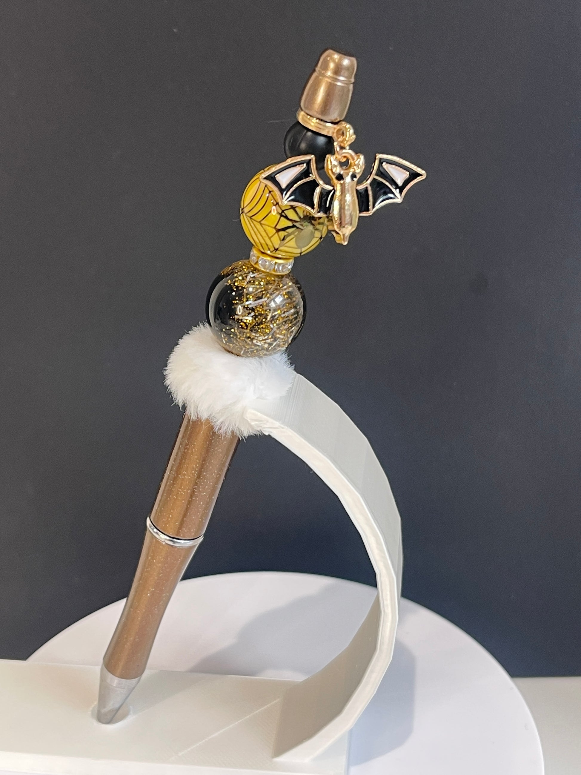 Decorative Halloween Beaded pen with black and gold beads and a white fluffy bead and a black and gold bat charm in front of a dark background
