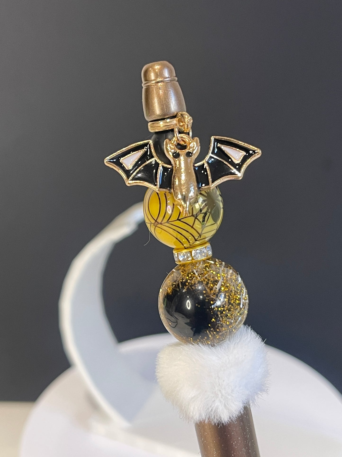 Close up of a decorative Halloween beaded pen with gold and black beads and a gold and black bat charm in front of a dark background.