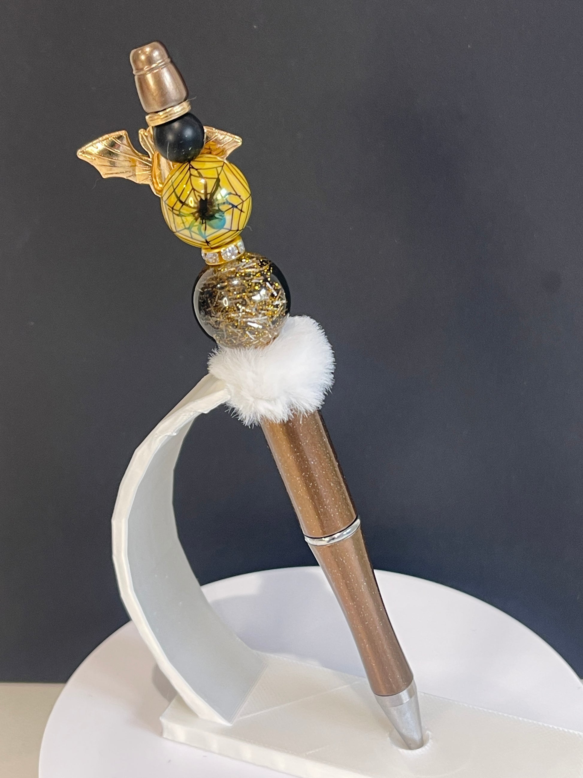 Decorative Halloween beaded pen with gold and black beads with an image of a spider and a white fluffy bead in front of a dark background
