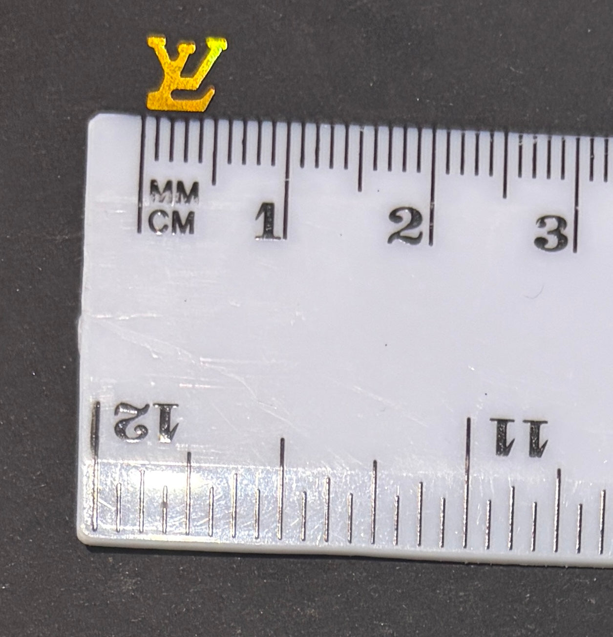 Single LV logo holographic gold glitter shape next to a white ruler on black background